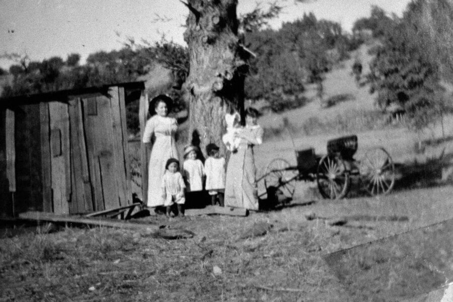 Two women and three small children stand in front of a large tree and a wooden shed in a rural landscape. An old-fashioned horse-drawn carriage is visible in the background. The image is black and white and slightly blurry.