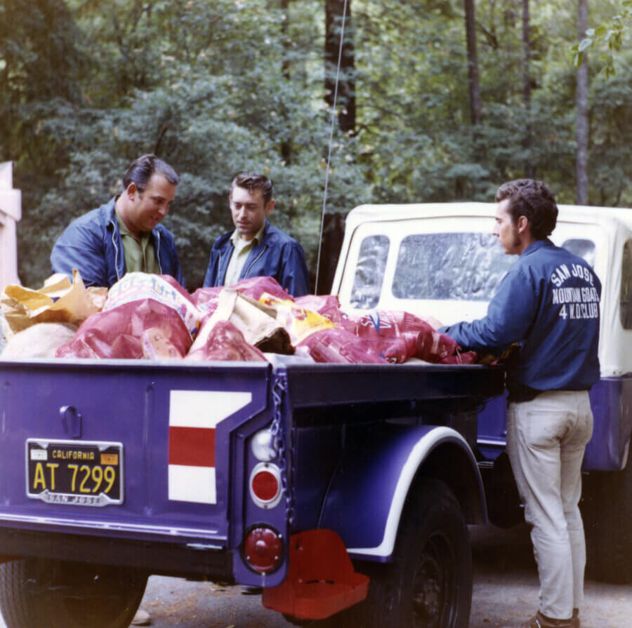 Three people in blue jackets stand around a truck filled with bags, possibly containing animal feed, in a wooded outdoor setting. The truck has a California license plate.